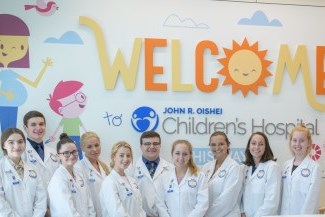 students in white lab coats in front of an Oshei Children's Hospital Welcome sign